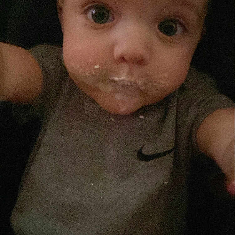 Neptune is registered to the contest to win money with this photo: baby, child, close_up, crumbs, curious, cute, expression, face, food, grey_clothing, indoor, infant, messy_face, nike_logo, person, portrait, selfie, shirt, wide_eyes, young_child