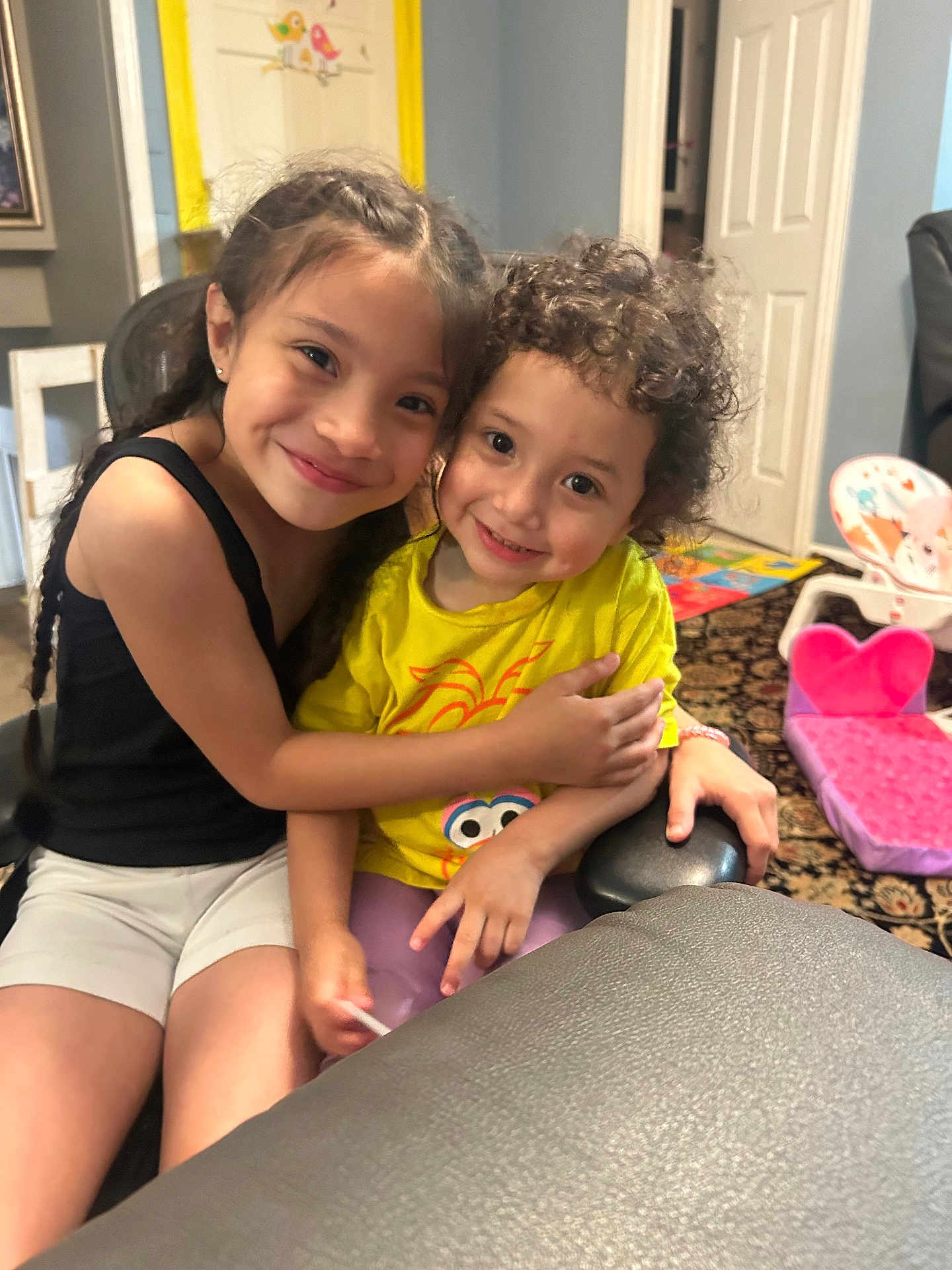 Josephine is registered to the contest to win money with this photo: child, children, smile, hug, indoor, toy, chair, heart_shaped_chair, yellow_shirt, black_top, curly_hair, braided_hair, happy, carpet, home, person, cute, sibling, playroom, friendly