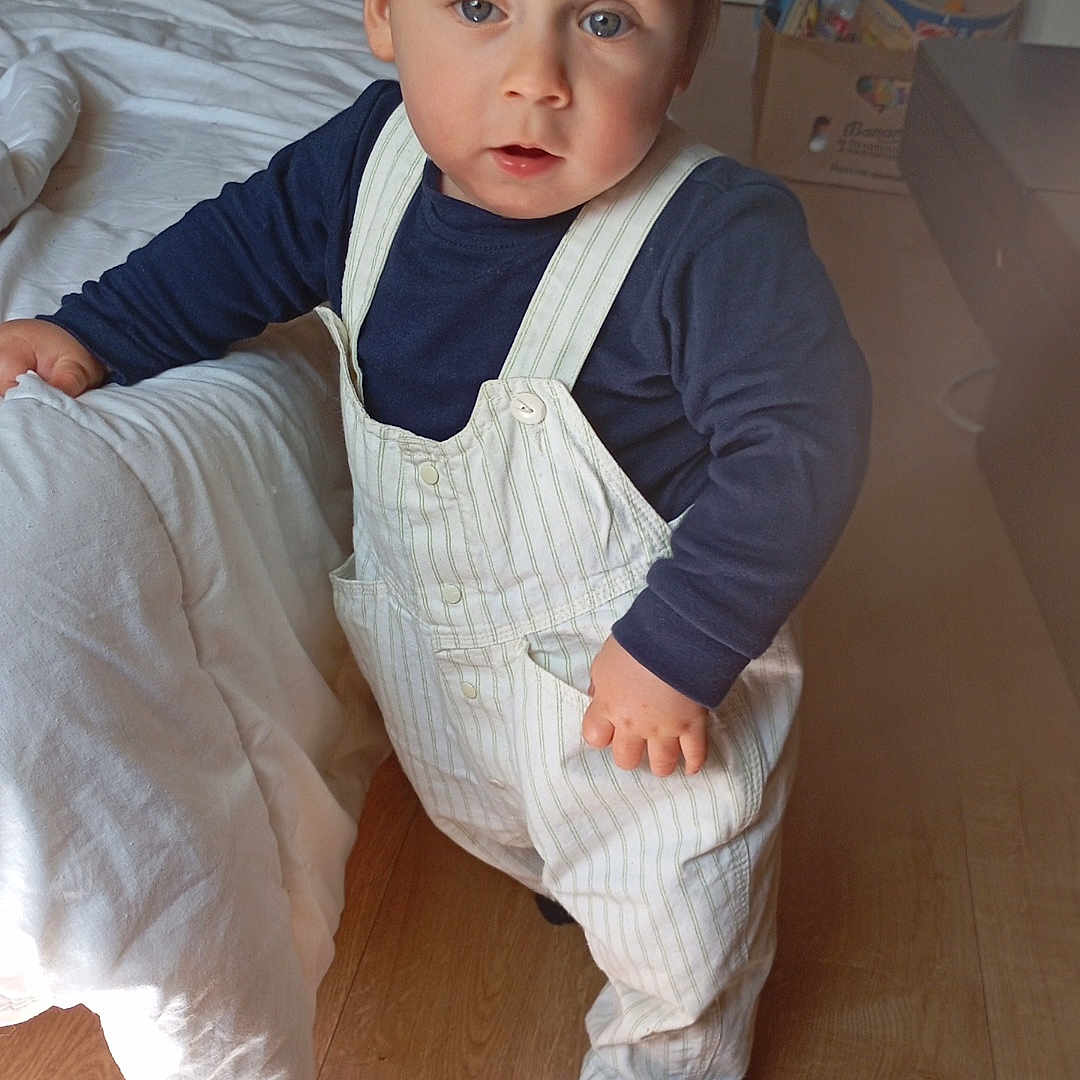 Léo participe au concours pour gagner de l'argent avec cette photo : baby, blanket, blue_eyes, casual_clothing, child, curious, floor, home, indoor, light_hair, long_sleeve_shirt, overalls, pale_skin, person, portrait, sock, standing, toddler, toy, wooden_floor