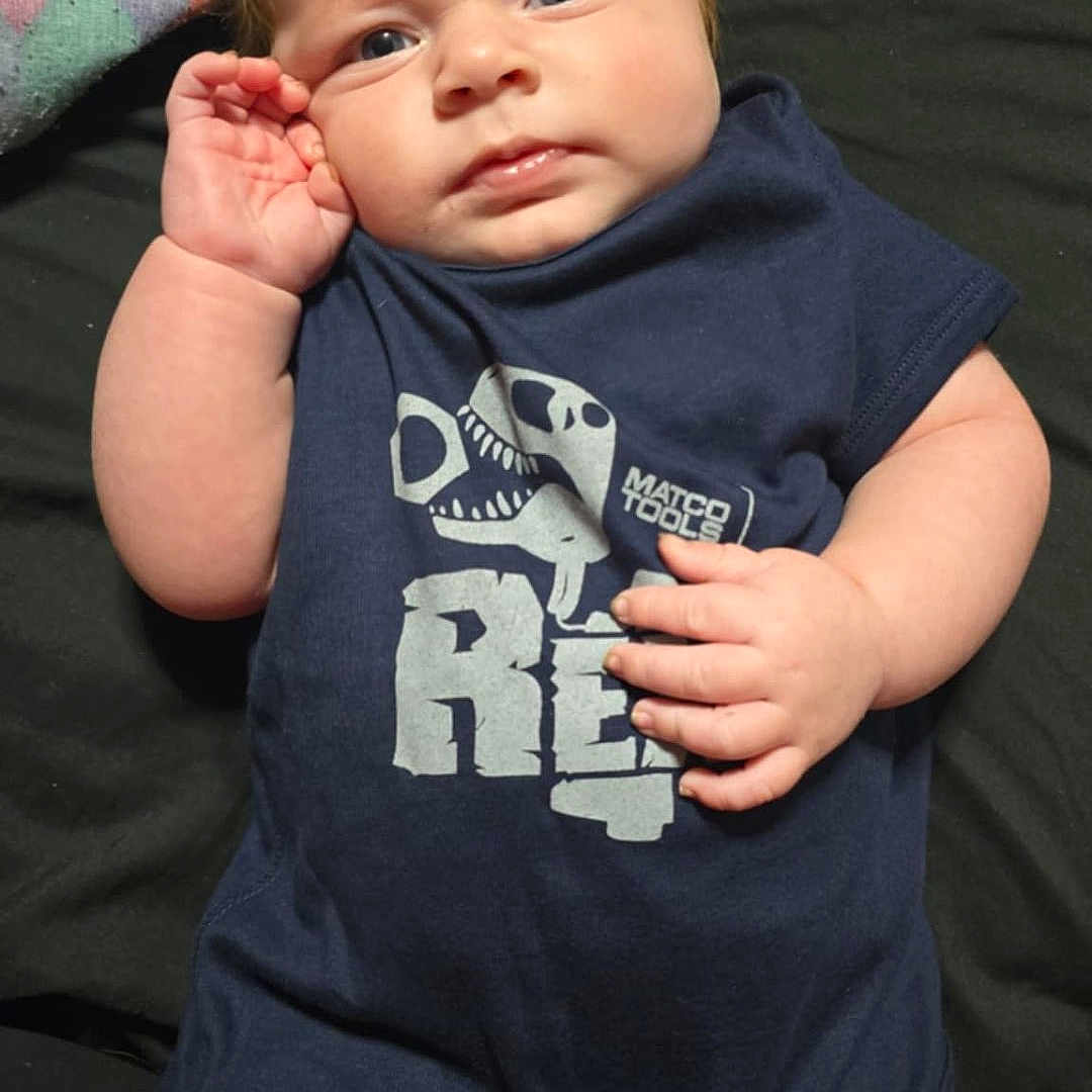 Kenneth joined the competition — help win amazing prizes! baby, blanket, blue_clothing, casual, chubby, curious_expression, dinosaur_graphic, eyes, face, fingers, hand, head, human, indoors, infant, lying_down, onesie, person, portrait, skin