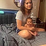 child, baby, bed, blanket, curly_hair, indoor, person, smiling, sitting, young_girl, furniture, mirror, wall_art, water_bottle, shorts, tshirt, skin, hand, foot, room