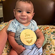 Isaac is registered to the contest to win money with this photo: baby, infant, smile, blue_onesie, wooden_sign, four_months_old, cushion, animal_print, blanket, chair, happy, child, portrait, indoors, cute, person, seated, holding, face, young