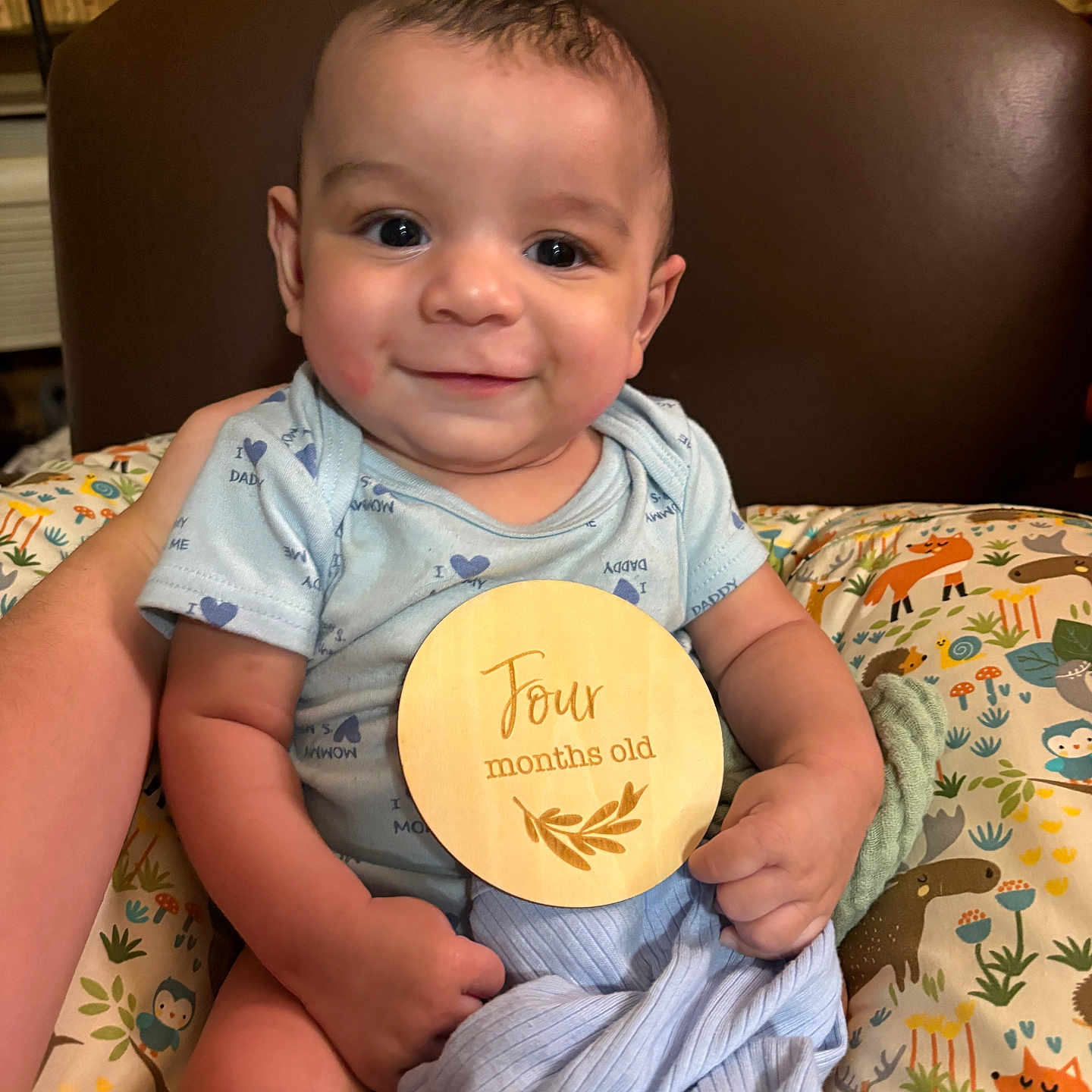 Isaac is registered to the contest to win money with this photo: baby, infant, smile, blue_onesie, wooden_sign, four_months_old, cushion, animal_print, blanket, chair, happy, child, portrait, indoors, cute, person, seated, holding, face, young