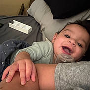 Isaac is registered to the contest to win money with this photo: baby, infant, child, smiling, happy, face, portrait, indoors, bed, bedroom, pillow, blanket, pacifier, clothing, gray_clothing, adult_arm, hand, family, lying_down, cozy