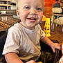 Luca is registered to the contest to win money with this photo: toddler, child, smiling, high_chair, food, nugget, restaurant, table, chair, white_shirt, camouflage_pants, happy, indoor, person, face, hand, wooden_table, background, sign, casual