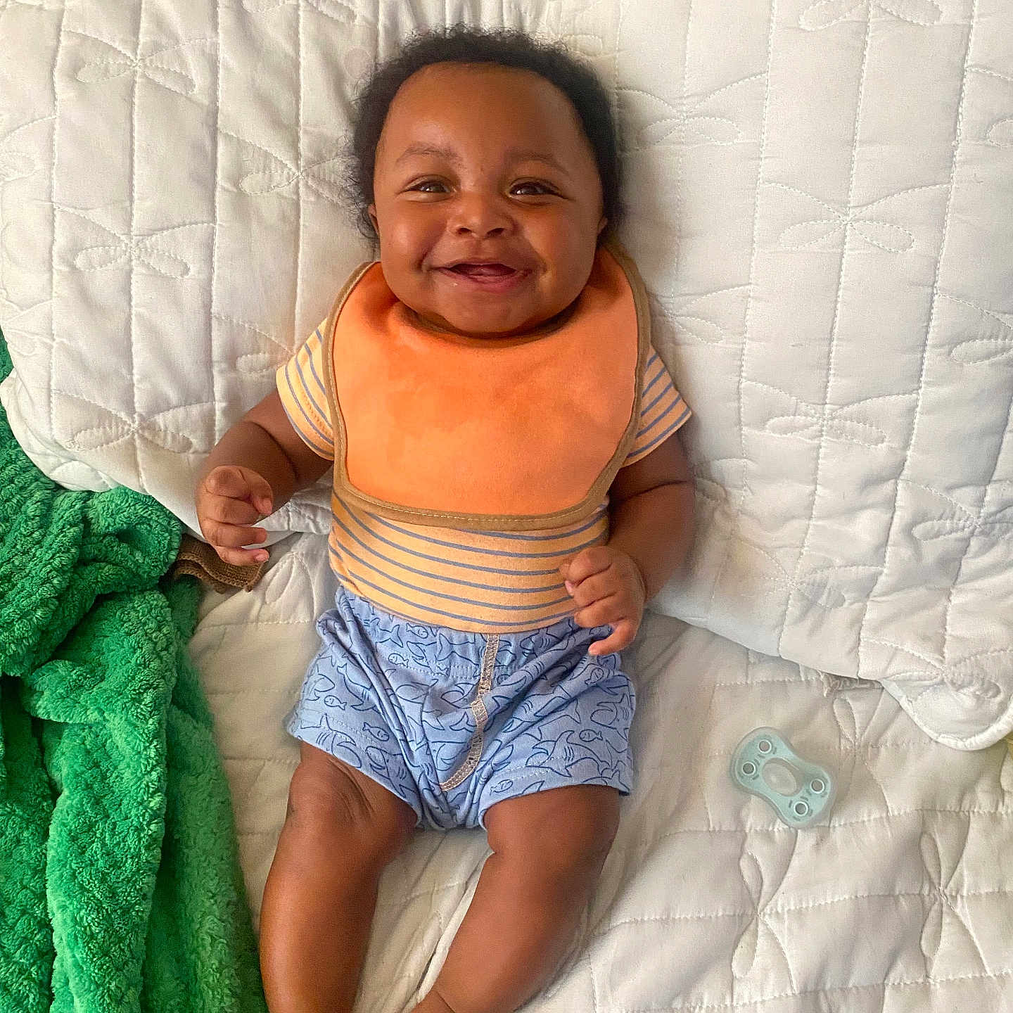Haissam is registered to the contest to win money with this photo: baby, bib, blanket, blue_shorts, child, cute, feet, green_towel, hand, happy, indoors, infant, legs, pacifier, person, quilted_bedspread, relaxed, skin, smiling, striped_shirt