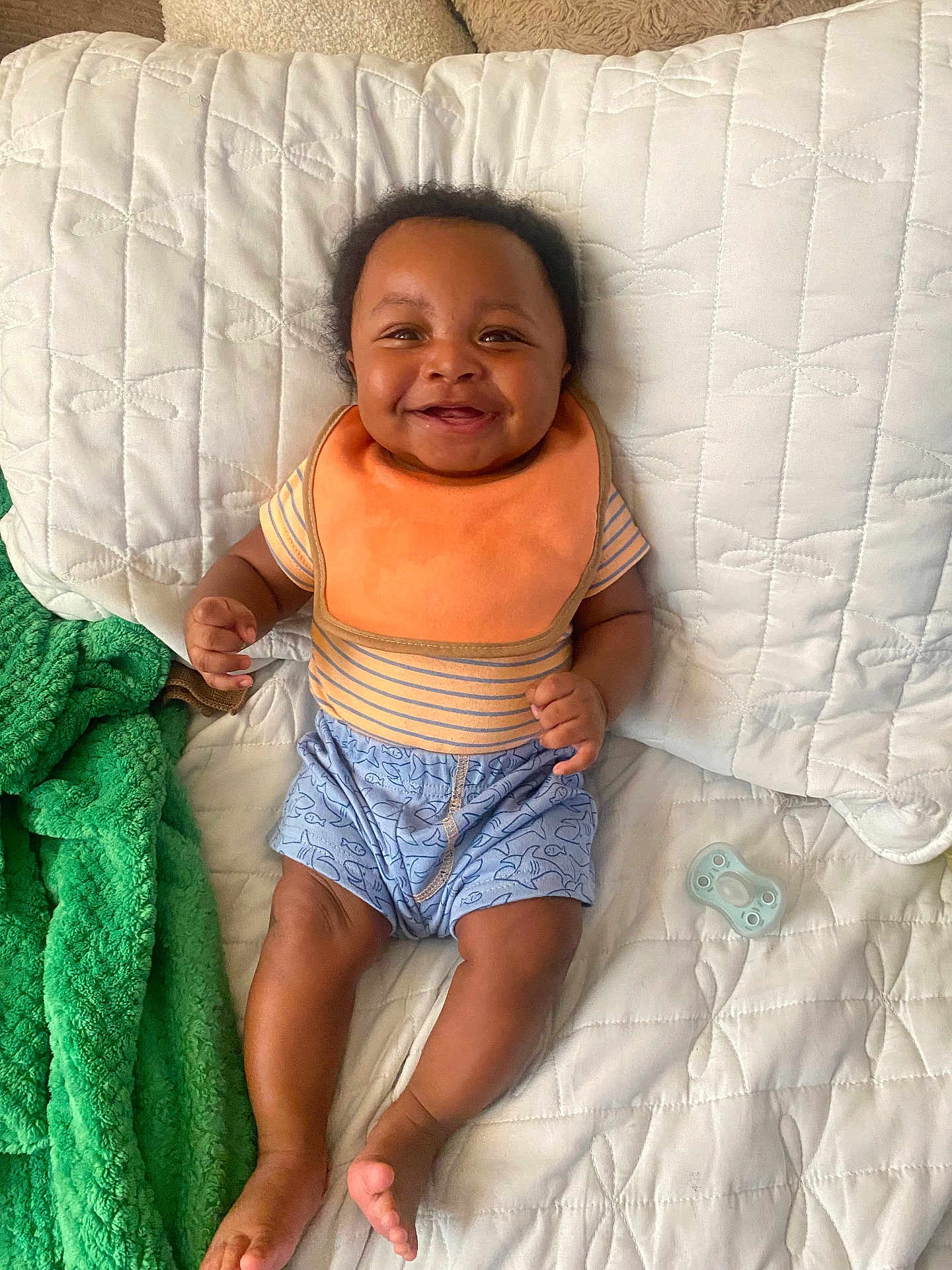 Haissam is registered to the contest to win money with this photo: baby, smiling, bib, striped_shirt, blue_shorts, pacifier, blanket, green_towel, quilted_bedspread, happy, infant, child, cute, relaxed, indoors, person, skin, legs, feet, hand