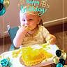 baby, birthday, cake, high_chair, food, smiling, celebration, party, decorations, balloons, roses, yellow_cake, face, child, messy, smile, indoor, table, hands, young_child