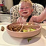 baby, background, black_curtain, cheeks, child, clothing, food, high_chair, indoor, kitchen, meat, messy_face, nose, person, plate, reaching, salad, seat, smiling, table