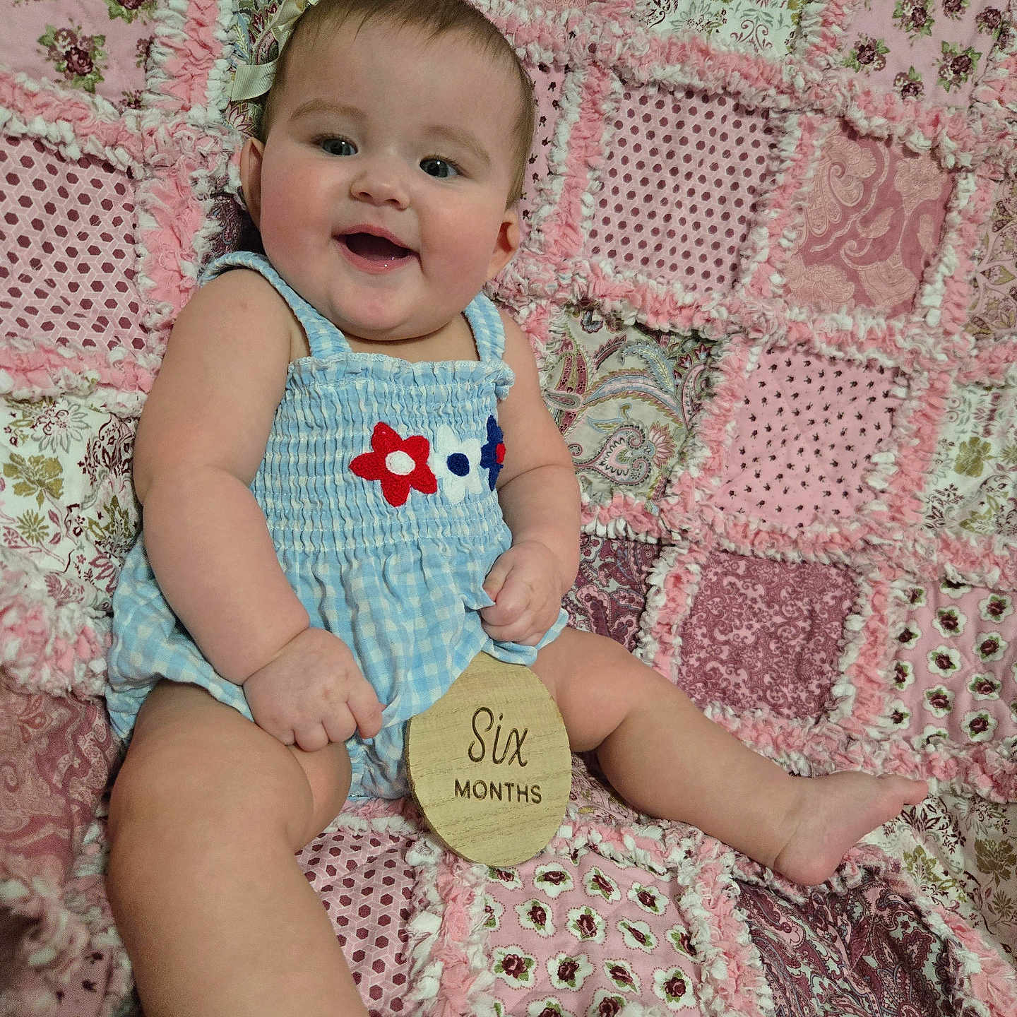 Lily joined the competition — help win amazing prizes! baby, child, infant, smiling, blue_clothing, flower_pattern, plush_quilt, pink, patchwork, sitting, milestone, wooden_plaque, happy, cute, portrait, indoors, person, skin, head, hand