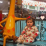 Kylie is registered to the contest to win money with this photo: toddler, child, toy_store, giraffe_statue, bench, holiday_pajamas, curly_hair, indoor, floor, sitting, festive, calendar_overlay, toys, shelf, red_bow, cute, playful, decor, lighting, portrait