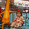 toddler, child, toy_store, giraffe_statue, bench, holiday_pajamas, curly_hair, indoor, floor, sitting, festive, calendar_overlay, toys, shelf, red_bow, cute, playful, decor, lighting, portrait
