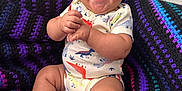 Demetrius is registered to the contest to win money with this photo: baby, laughing, smiling, infant, knitted_blanket, purple, black, blue, pink, onesie, dinosaur_print, sitting, happy, cute, indoor, child, person, soft_texture, cozy, small_feet