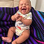 baby, laughing, smiling, infant, knitted_blanket, purple, black, blue, pink, onesie, dinosaur_print, sitting, happy, cute, indoor, child, person, soft_texture, cozy, small_feet