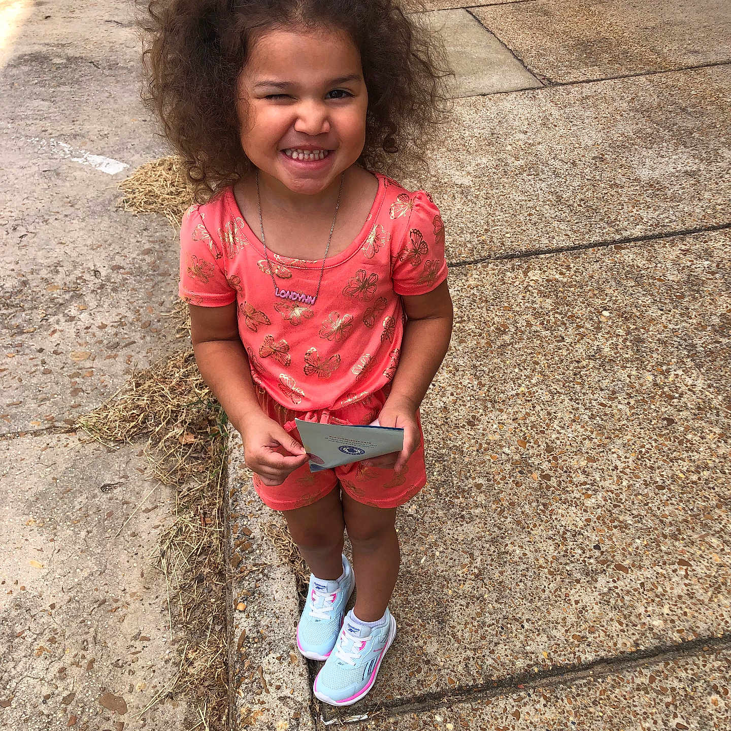 Londynn is registered to the contest to win money with this photo: butterfly_pattern, casual_clothing, child, concrete, coral_outfit, curly_hair, daylight, girl, happy, holding_object, outdoor, playful, portrait, scrunchie, sidewalk, smiling, standing, sunny, white_sneakers, winking