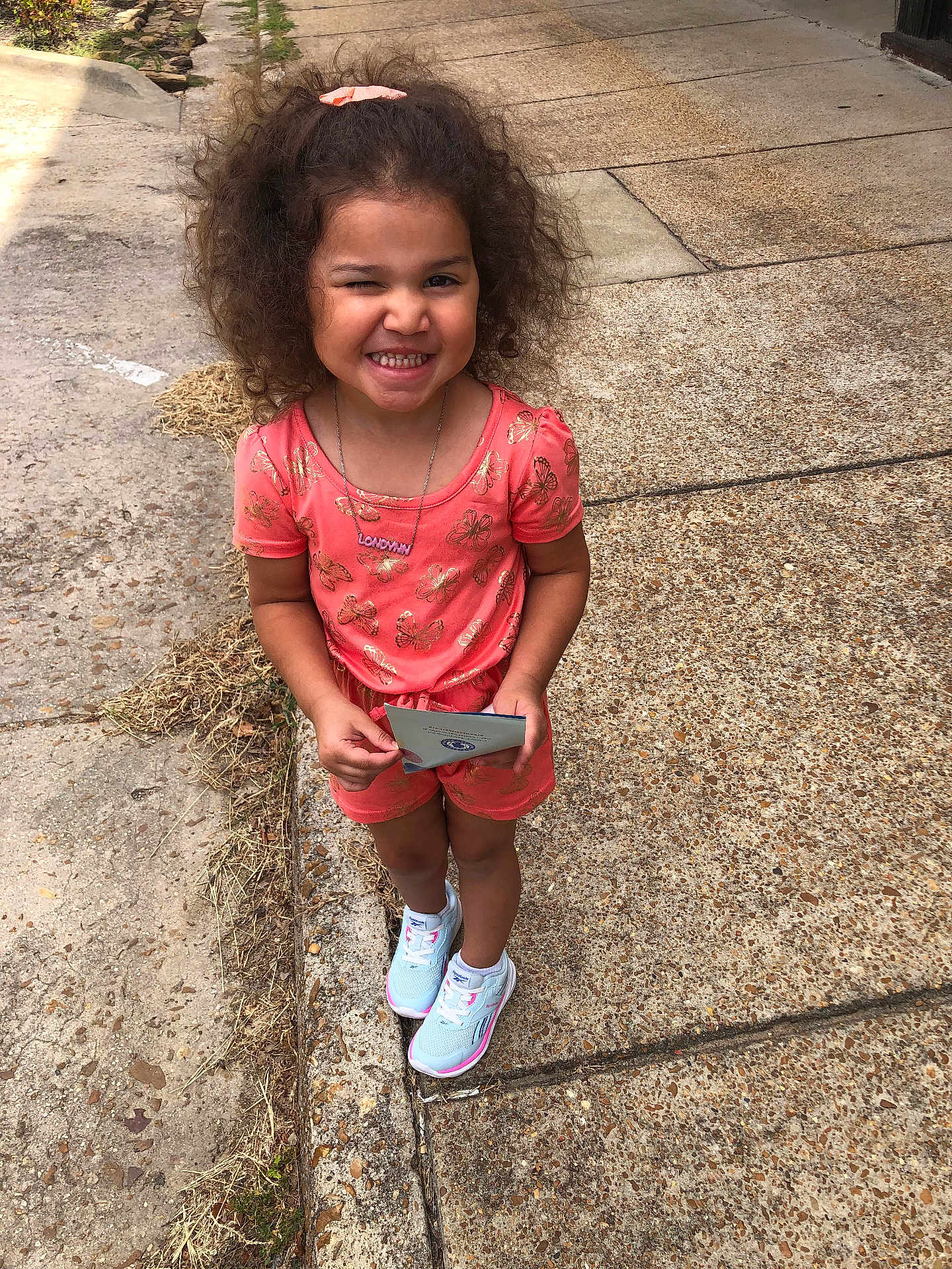Londynn is registered to the contest to win money with this photo: child, girl, smiling, winking, curly_hair, coral_outfit, butterfly_pattern, white_sneakers, sidewalk, concrete, outdoor, sunny, playful, holding_object, scrunchie, happy, standing, casual_clothing, portrait, daylight