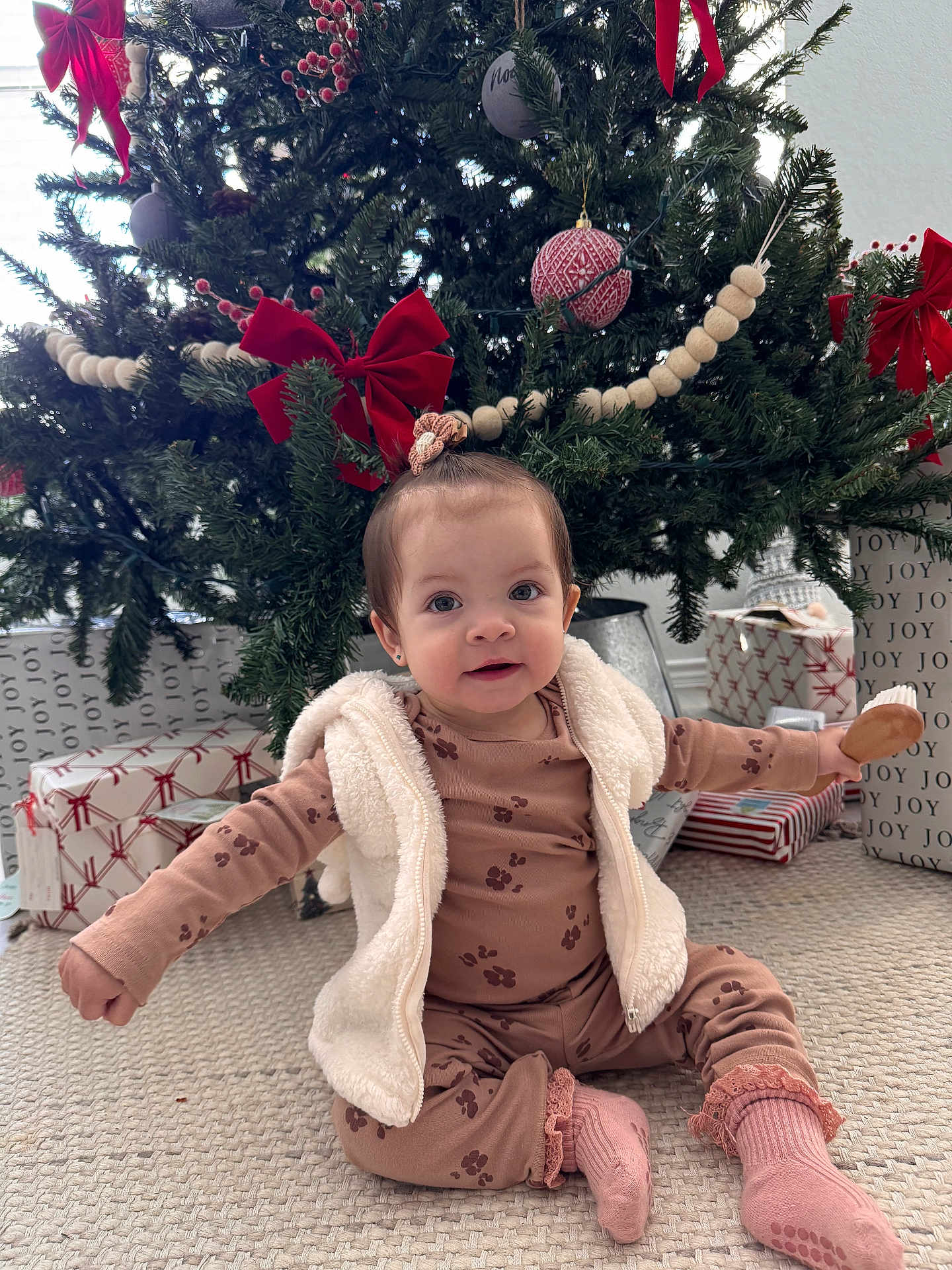 Maríaelisa is registered to the contest to win money with this photo: baby, child, christmas_tree, holiday, presents, gift_wrap, red_bow, carpet, indoor, festive, decorations, vest, pajamas, socks, cute, smiling, sitting, toy, holiday_season, home