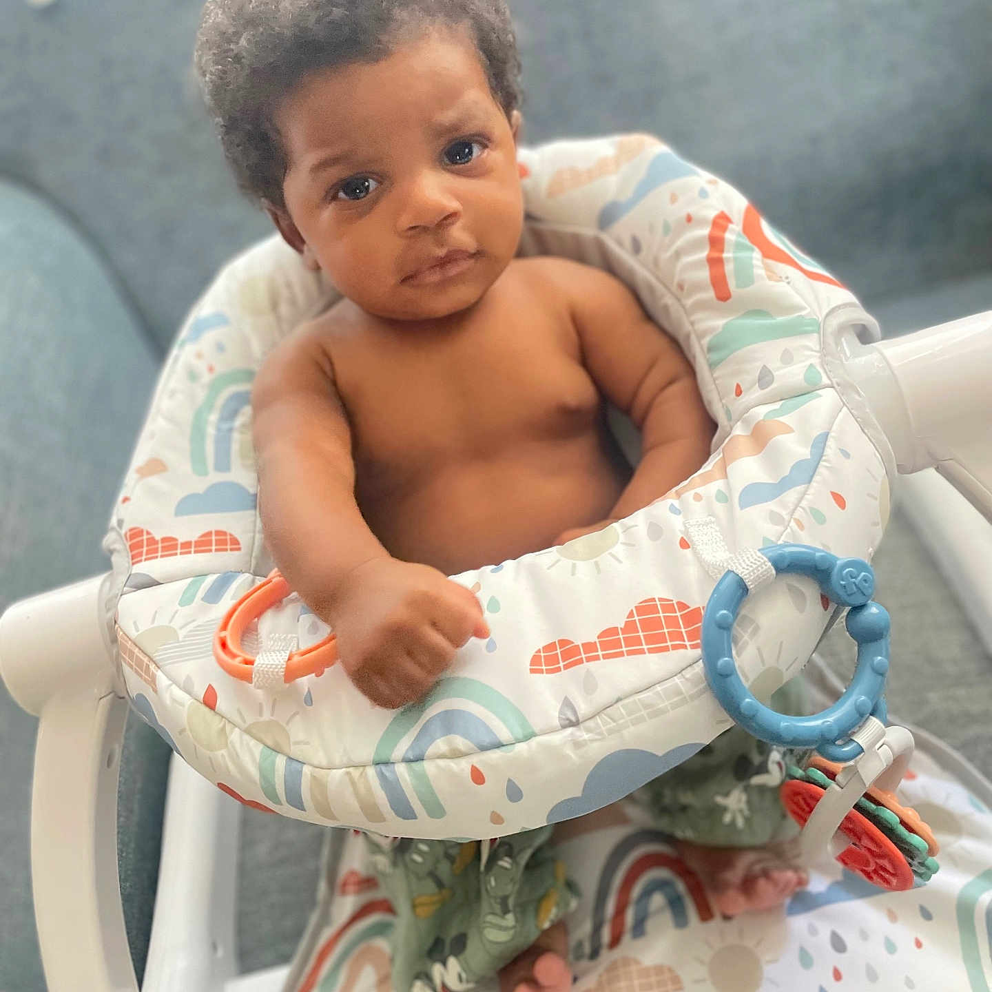 Eleven is registered to the contest to win money with this photo: baby, infant, child, curly_hair, bare_chest, baby_seat, rainbow_pattern, colorful, toy_ring, foot, hand, indoor, couch, fabric, cute, looking_at_camera, portrait, playful, small_person, furniture