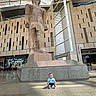 Adham is registered to the contest to win money with this photo: baby, child, statue, museum, indoor, floor, ancient_art, stone, modern_architecture, curious, blue_shirt, large, contrast, sitting, light, shadow, public_space, tourist, monument, art