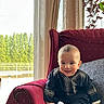 baby, child, smiling, sitting, armchair, red_chair, traditional_clothing, indoors, window, curtains, cushion, plush_furniture, footwear, white_socks, happy, portrait, person, home, greenery, daylight