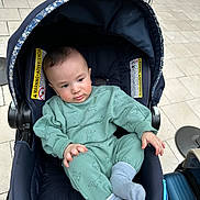 Adham is registered to the contest to win money with this photo: baby, child, stroller, infant, clothing, green_outfit, socks, person, floor, tile, luggage, footwear, indoor, warning_label, navy_blue, cute, seated, curious, hands, head