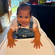 Melany is registered to the contest to win money with this photo: baby, bow, child, daylight, denim_overalls, doorway, face, floral_shirt, furniture, hands, happy, high_chair, home, indoor, portrait, sitting, smiling, television, tray, window
