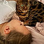 cat, bengal_cat, green_eyes, child, person, bed, blanket, pillow, braid, fur, whiskers, striped_pattern, cozy, cuddle, resting, portrait, indoor, close_up, sleepy, pet