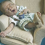 Wyatt is registered to the contest to win money with this photo: baby, doll, glasses, sleeping, armchair, miniature, slippers, latte, coffee, latte_art, plaid_clothing, beige, blue_background, rug, hand, foot, cute, indoors, toy, relaxing