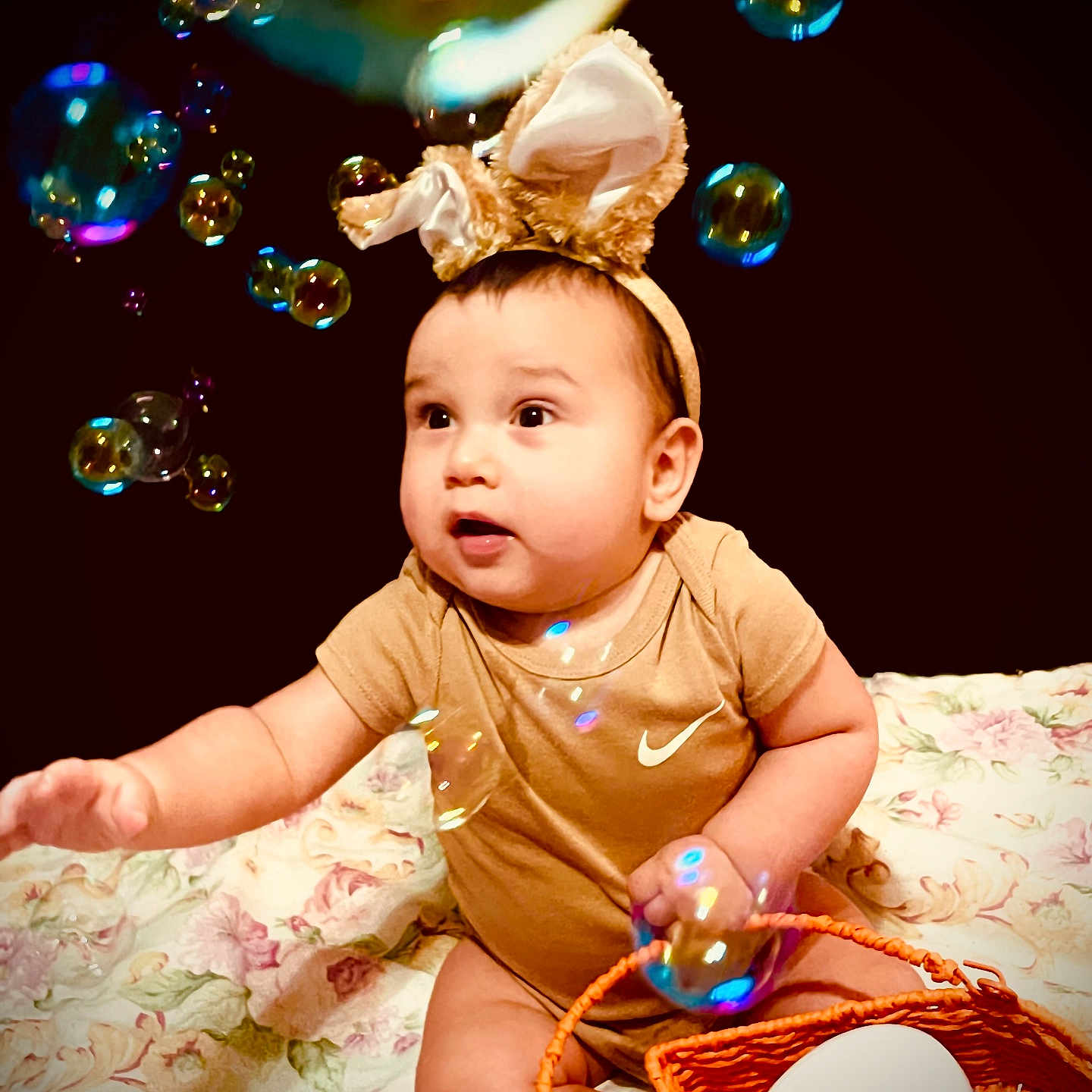 Barric is registered to the contest to win money with this photo: baby, child, bubbles, bunny_ears, basket, blanket, container, floral_pattern, curious, indoor, cute, infant, sitting, reaching, playful, toy, face, headband, clothing, expression