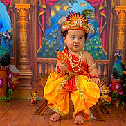 Ram joined the competition — help win amazing prizes! toddler, child, traditional_clothing, orange_dress, headgear, peacock, decorations, wooden_floor, jewelry, cultural, festive, portrait, indoor, colorful, smiling, small_stool, ornate_background, toy, statues, celebration
