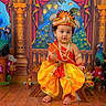 toddler, child, traditional_clothing, orange_dress, headgear, peacock, decorations, wooden_floor, jewelry, cultural, festive, portrait, indoor, colorful, smiling, small_stool, ornate_background, toy, statues, celebration