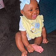 Miangel is registered to the contest to win money with this photo: baby, child, smiling, sitting, couch, headband, bow, yellow_bib, foot, hand, happy, indoors, clothing, person, portrait, cute, infant, accessory, jewelry, skin