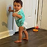toddler, child, walking, standing, indoors, portrait, face, short_hair, turquoise_shirt, diaper, barefoot, wall, door, floor, toy, red_toy, orange_ring, home, shadow, candid