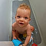 toddler, child, bathtub, water, toy, blue_cup, smile, happy, bath_toys, red_star_toy, yellow_toy, bathmat, naked, curly_hair, indoors, play, sitting, face, person, cute