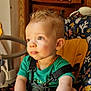 toddler, child, high_chair, green_shirt, amber_necklace, drool, curly_hair, indoor, wooden_furniture, colorful_pattern, seatbelt, baby, portrait, looking_away, cute, young_child, sitting, daylight, expression, face