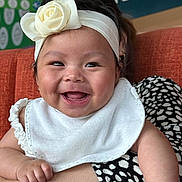 Lauren is registered to the contest to win money with this photo: baby, smile, headband, flower, bib, arm, person, clothing, couch, orange, polka_dot, black_and_white, happy, indoor, holding, face, skin, child, background, portrait
