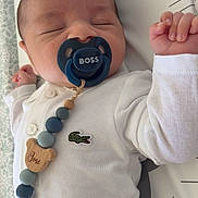 Joseph participe au concours pour gagner de l'argent avec cette photo : baby, sleeping, pacifier, white_shirt, beaded_pacifier_clip, crocodile_logo, infant, cute, child, newborn, hand, face, closed_eyes, lying_down, bed, blanket, clothing, blue, wooden_beads, cute_accessory