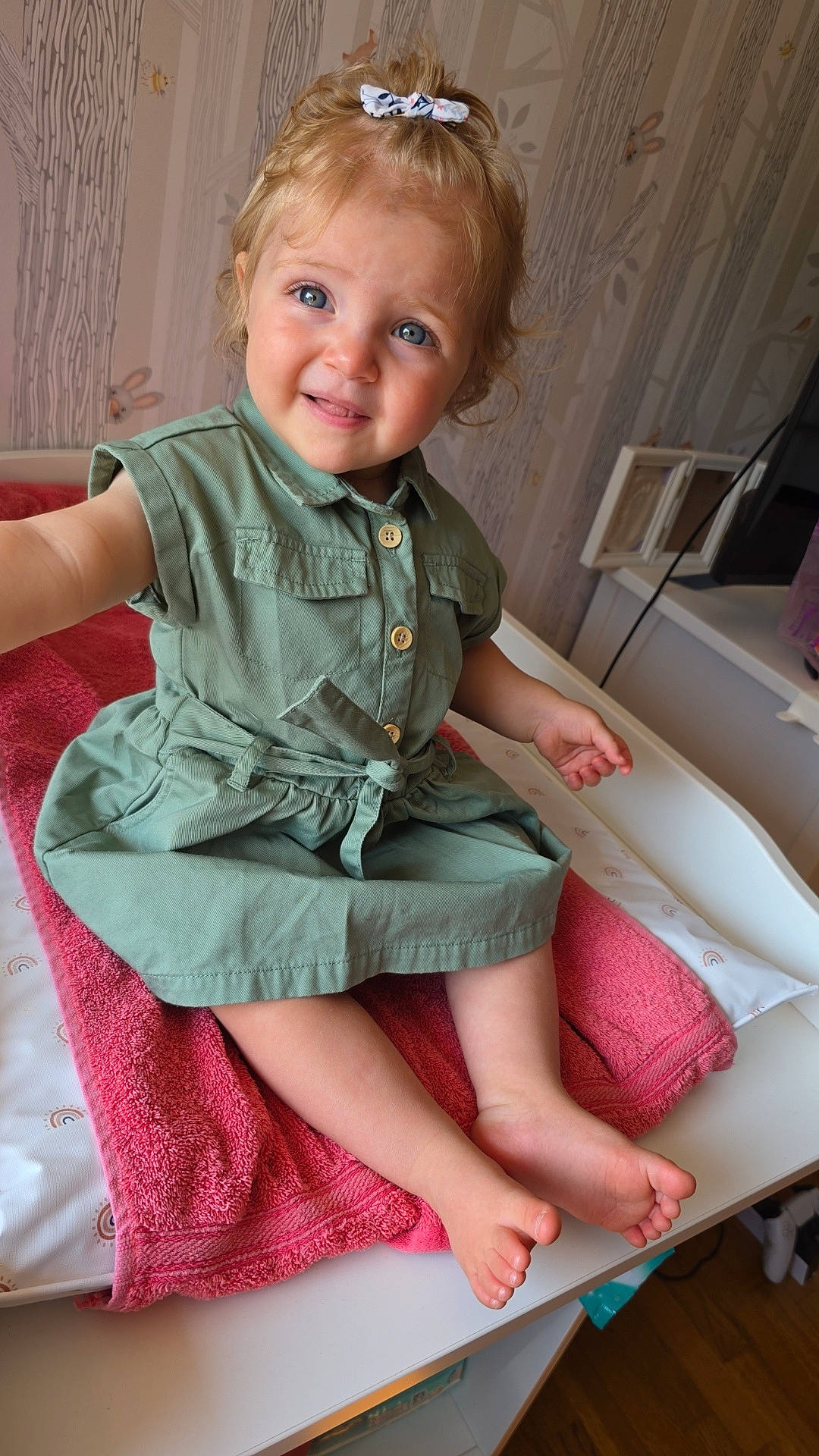 baby, barefoot, bee, blue_eyes, bow, bunny, changing_table, child, curly_hair, cute, furniture, green_dress, indoor, pink_towel, portrait, sitting, smile, toddler, wallpaper, wood_floor