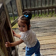 Nevaeh is registered to the contest to win money with this photo: toddler, child, outdoor, wooden_deck, railing, curious, blue_hair_tie, jeans, white_top, standing, portrait, daylight, young_child, casual_clothing, baby, person, exploring, natural_light, looking_back, expression