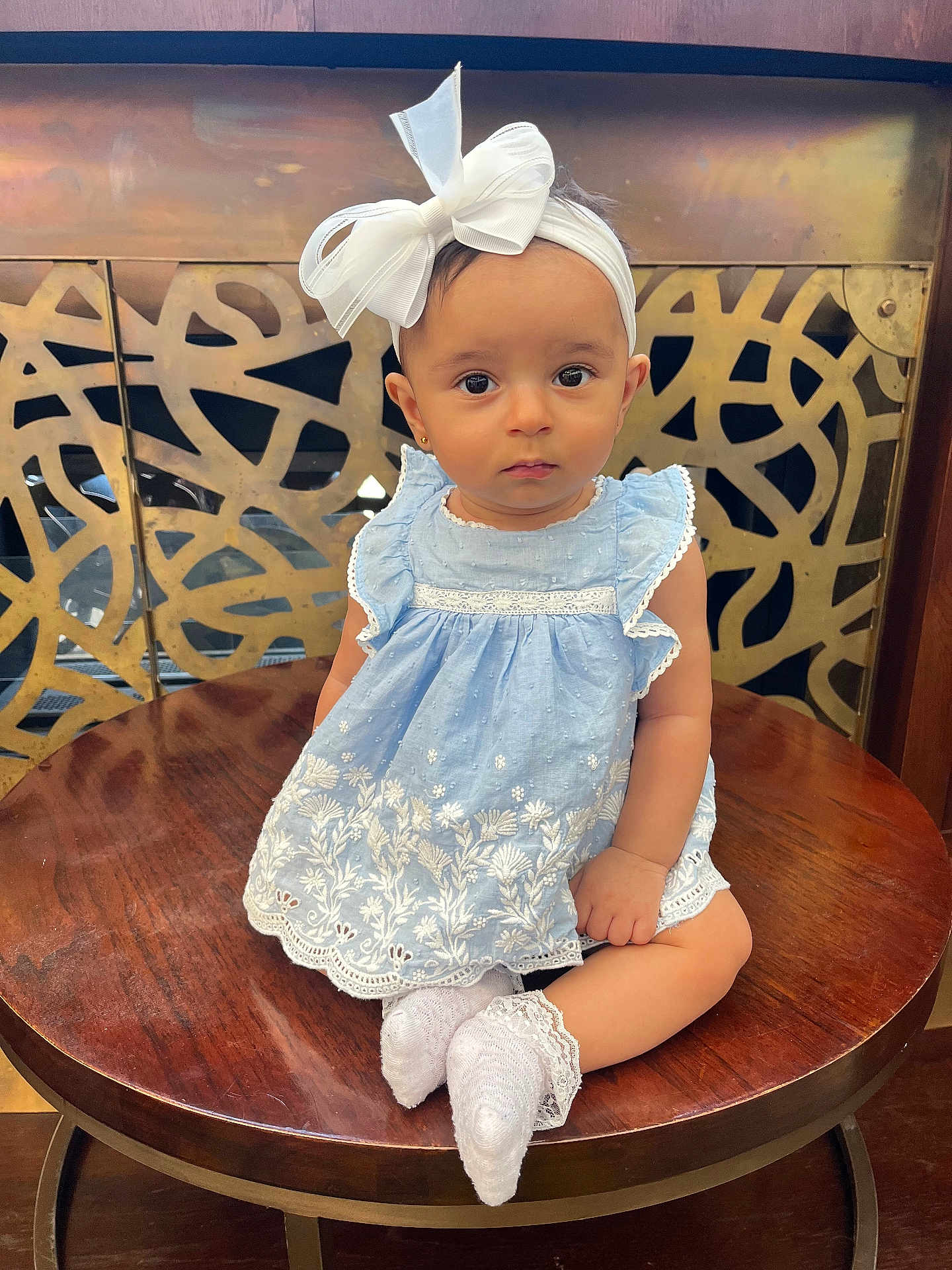 Mahratie is registered to the contest to win money with this photo: baby, child, girl, blue_dress, headband, bow, white_socks, wooden_table, decorative_background, face, portrait, sitting, cute, indoors, young_child, fashion, accessory, expression, eye_contact, closeup