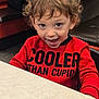 child, toddler, smile, happy, red_shirt, text_on_clothing, curly_hair, portrait, indoor, restaurant, table, booth_seating, eyes, playful, cute, dining, casual_clothing, person, expressive_face, young_child