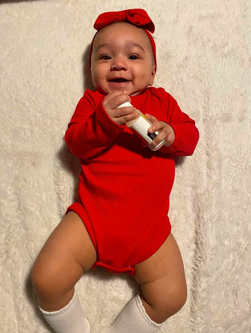 Makayla is registered to the contest to win money with this photo: baby, infant, red_onesie, headband, smiling, happy, holding_object, lying_down, white_blanket, soft_texture, cute, child, sock, skin, portrait, indoors, one_person, toddler, hands, face