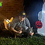 Crash is registered to the contest to win money with this photo: person, dog, small_dog, pet_owner, hat, cap, grass, brick_wall, ghost_decoration, pumpkin_decoration, jack_o_lantern, inflatable, night, outdoor, costume, crouching, portrait, illumination, yard, halloween
