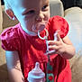 Miranda is registered to the contest to win money with this photo: toddler, child, pacifier, bottle, red_dress, indoor, couch, seated, curious_expression, blonde_hair, baby_accessory, patterned_pants, hand, face, person, young_child, home_interior, furniture, casual_clothing, soft_lighting