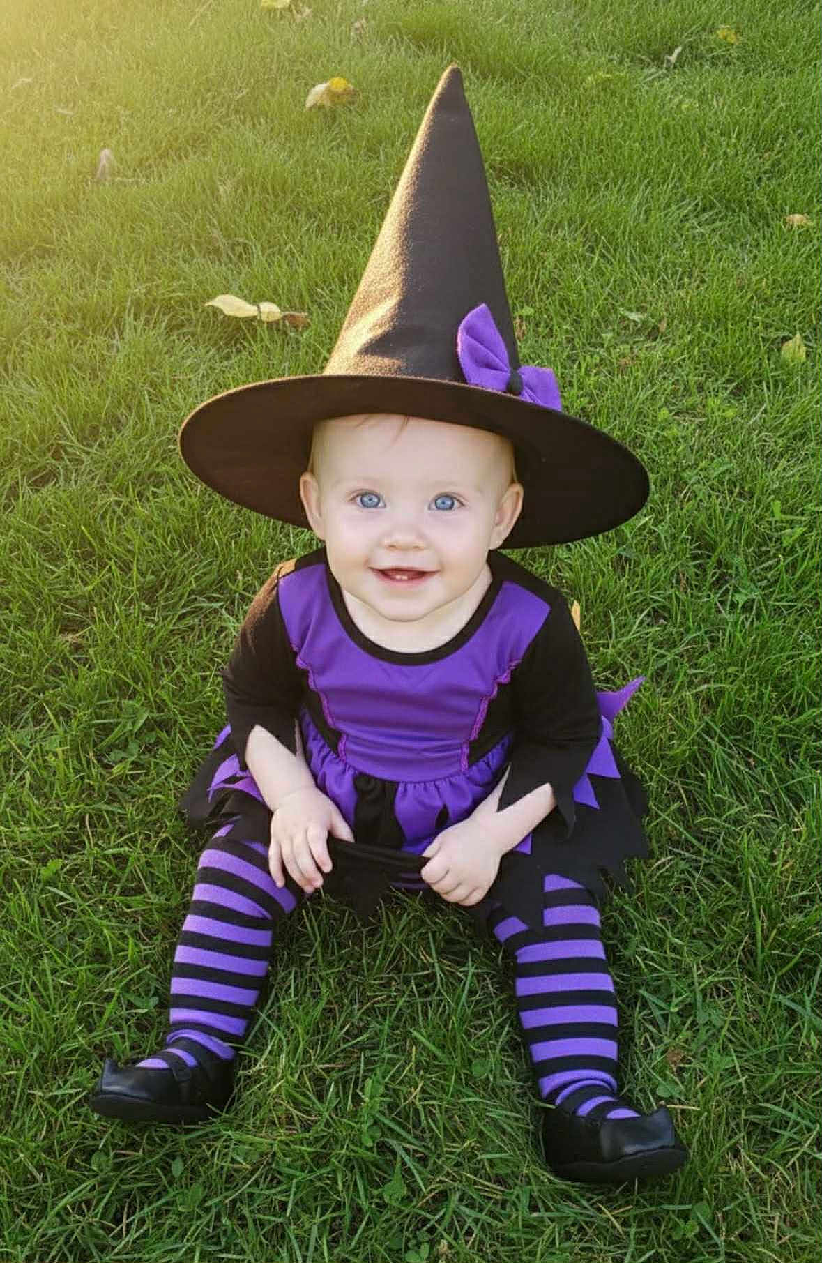 Miranda joined the competition — help win amazing prizes! baby, child, witch_costume, hat, purple, black, striped_tights, grass, outdoor, smile, blue_eyes, sunlight, costume, sitting, cute, happy, playful, portrait, holiday, festive