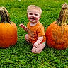 William is registered to the contest to win money with this photo: baby, child, pumpkin, grass, outdoor, smile, orange_shirt, fall, autumn, nature, greenery, cute, squatting, happy, person, seasonal, harvest, garden, young_child, playful