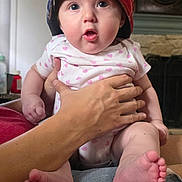 Cooper is registered to the contest to win money with this photo: baby, child, infant, hat, red_cap, onesie, hand, indoor, person, face, feet, jeans, expression, surprised, holding, skin, fingers, door, fireplace, home
