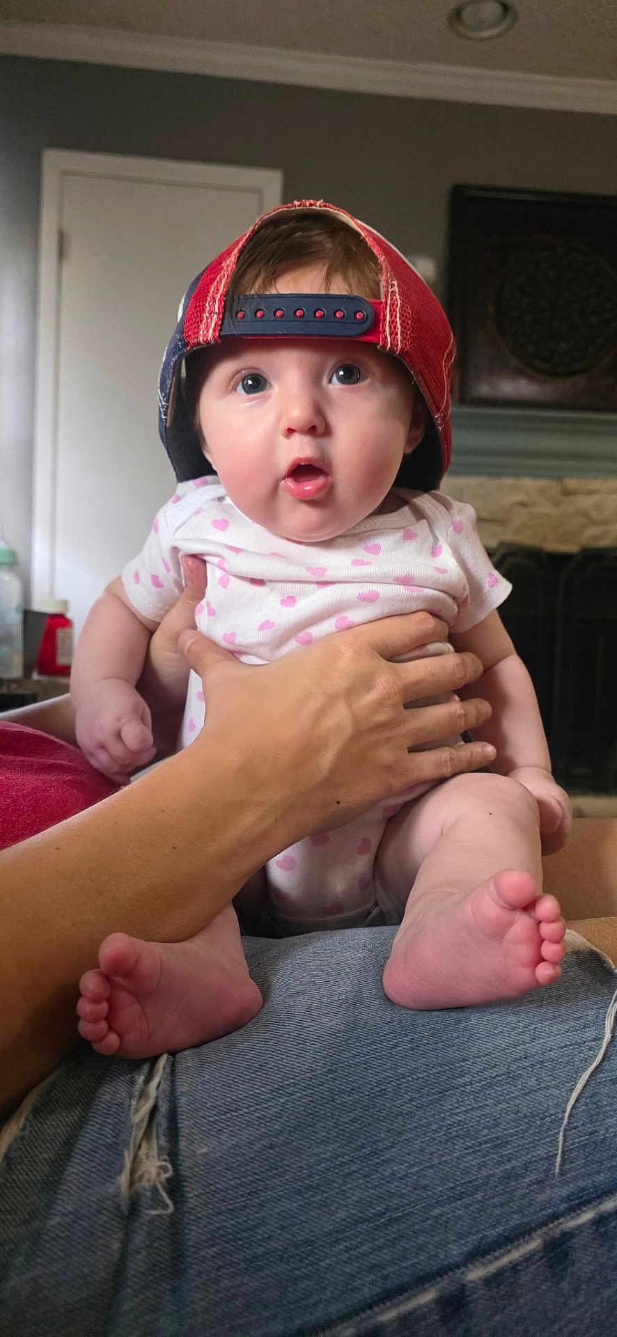 Cooper is registered to the contest to win money with this photo: baby, child, infant, hat, red_cap, onesie, hand, indoor, person, face, feet, jeans, expression, surprised, holding, skin, fingers, door, fireplace, home