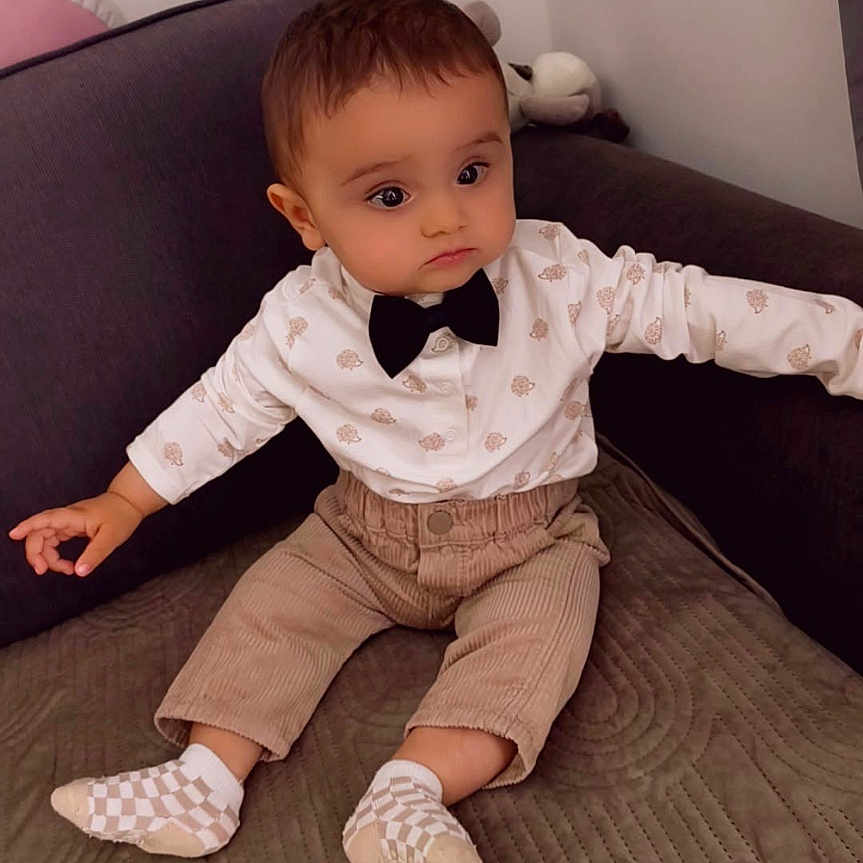 Kristian participe au concours pour gagner de l'argent avec cette photo : baby, beige_pants, bow_tie, casual_clothing, child, couch, curious, cute, expression, furniture, hands_out, home, indoor, portrait, seated, socks, soft_lighting, toddler, white_shirt, young_child