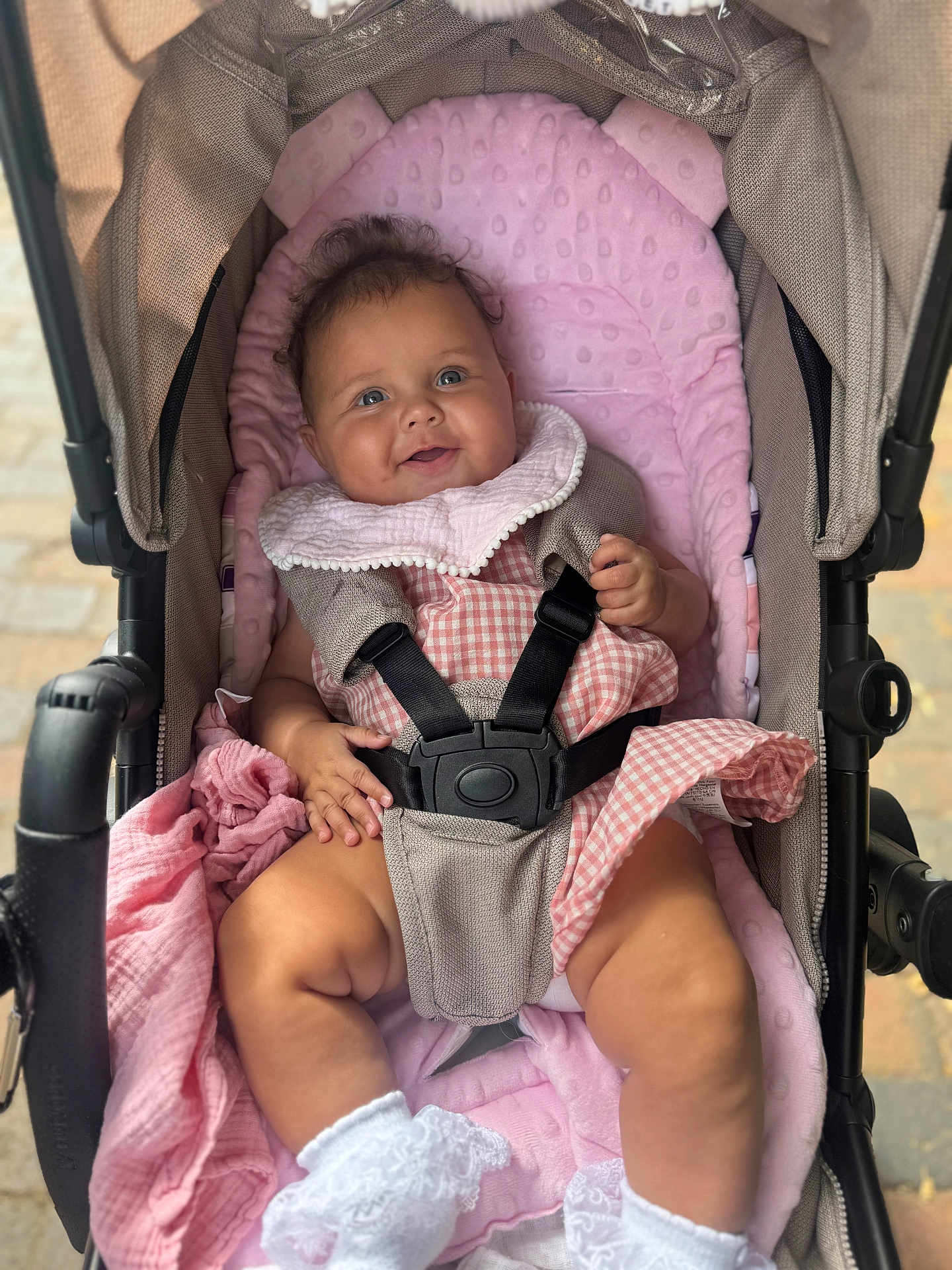 Ada-Violet is registered to the contest to win money with this photo: baby, stroller, pink_blanket, gingham_dress, white_socks, smiling, child, seatbelt, curly_hair, outdoor, happy, cute, infant, baby_clothing, footwear, baby_toys, fabric, baby_bib, soft_texture, baby_care